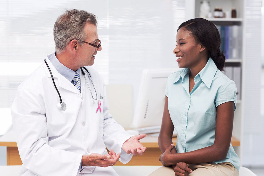 Male doctor with breast cancer pin is speaking with smiling female patient inside doctor's office