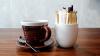 A coffee cup and spoon sit next to a cup full of sweeteners and sugar packets