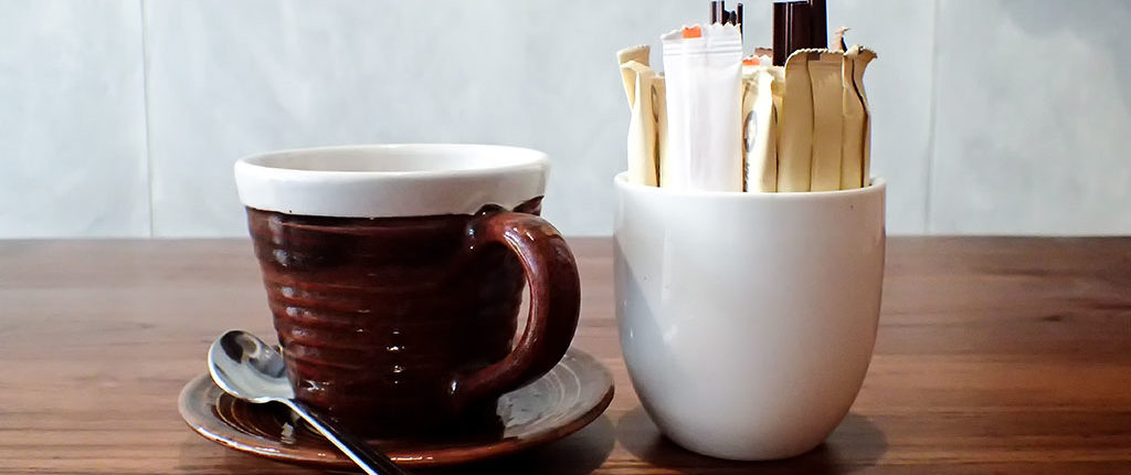 A dark brown saucer and matching mug with a white rim sit next to a white cup of sweetener packets