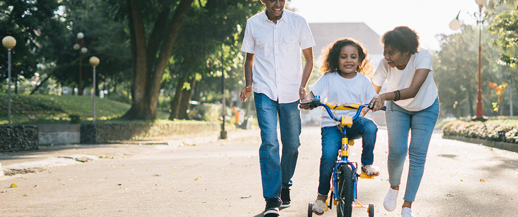 A man and woman walk down the street with a girl on a bicycle with training wheels