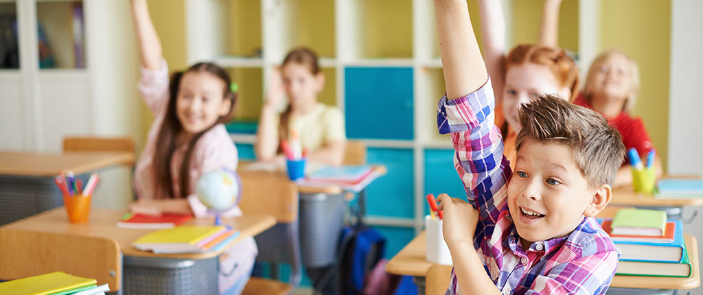 Part of a classroom, showing five adolescent children, all eagerly raising their hands