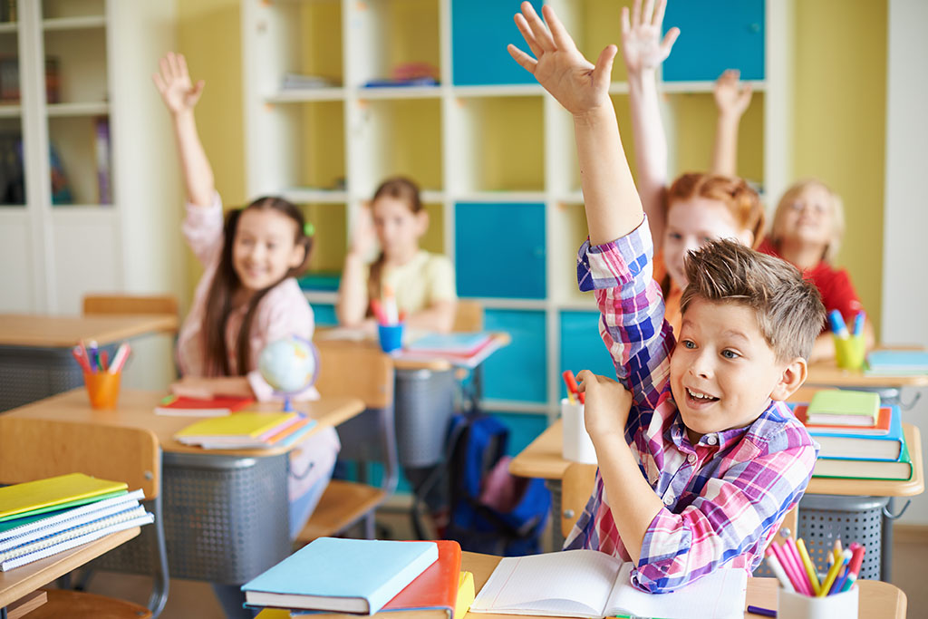 Part of a classroom, showing five adolescent children, all eagerly raising their hands