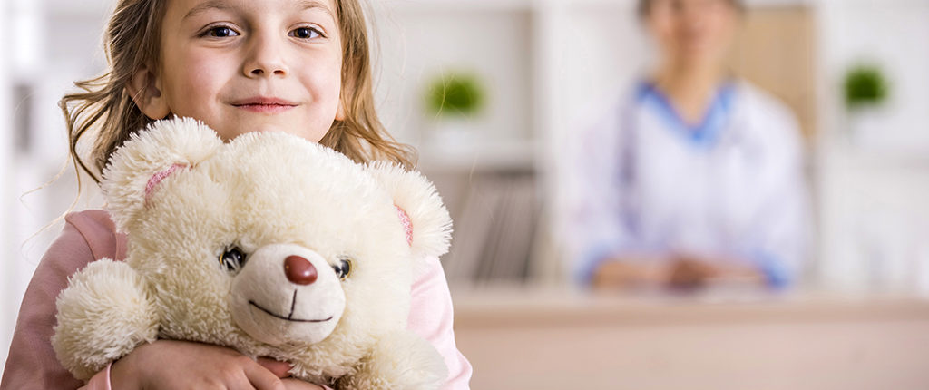 A young girl smiles while holding a stuffed animal. A woman is blurred in the background