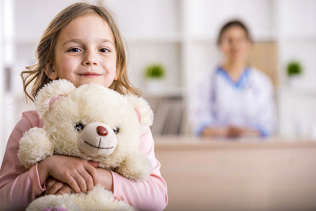 A young girl smiles while holding a stuffed animal. A woman is blurred in the background