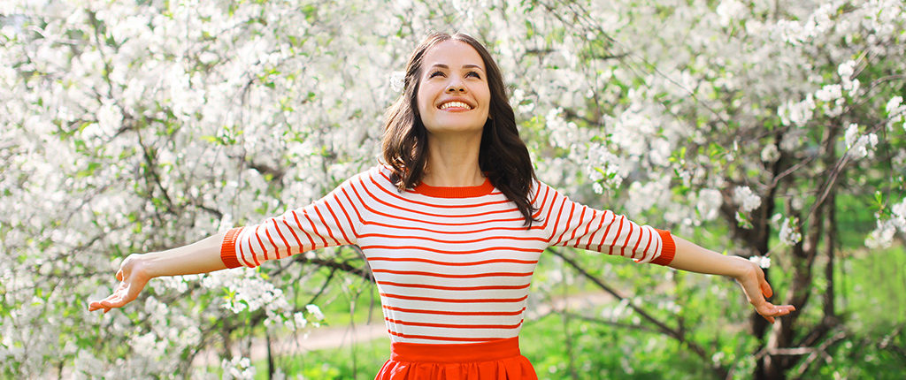A woman in a red and white shirt and red skirt stands smiling in an orchard