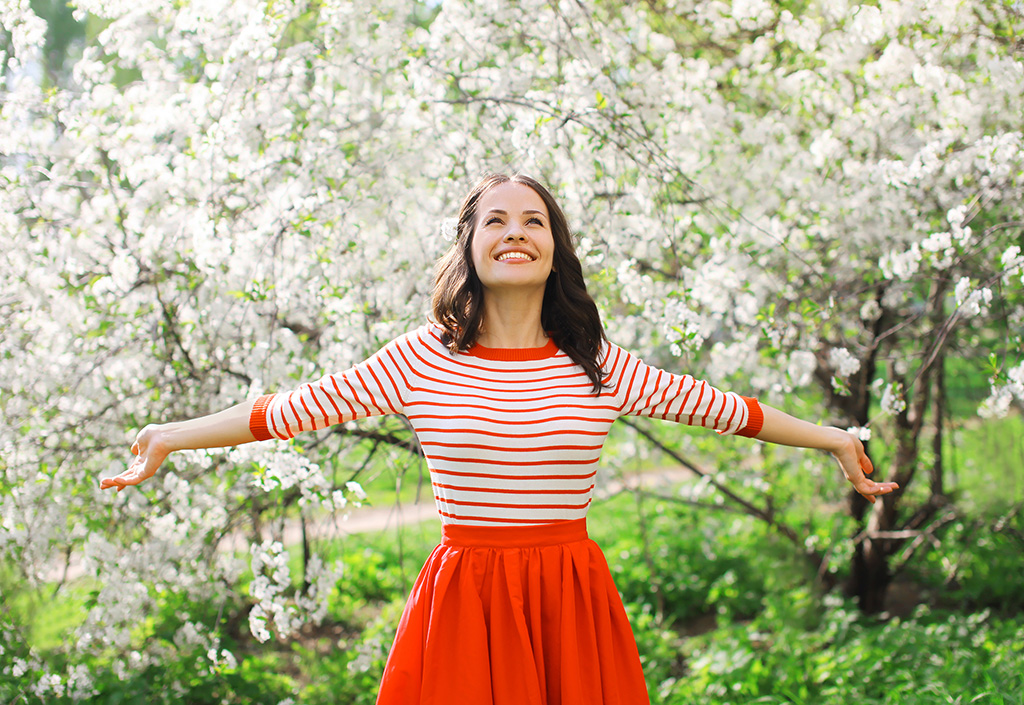 A woman in a red and white shirt and red skirt stands smiling in an orchard