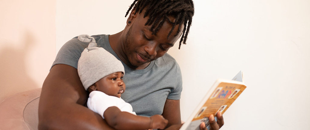 A man holds a baby in one arm and a book in the other, he is looking at the baby