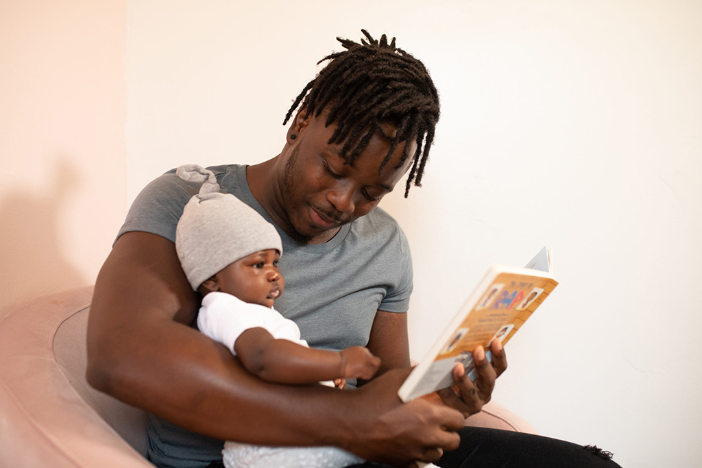 A man holds a baby in one arm and a book in the other, he is looking at the baby