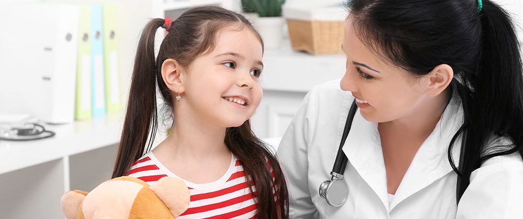 A woman in a lab coat holds a girl in a red and white shirt holding a stuffed animal