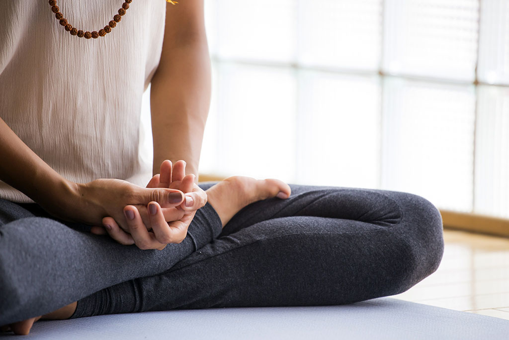 A woman sits in half lotus position on a yoga mat, her face is cropped out of the photo