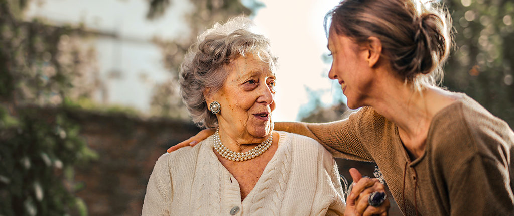 A younger woman has her arm around an older woman's shoulders, they are looking at each other