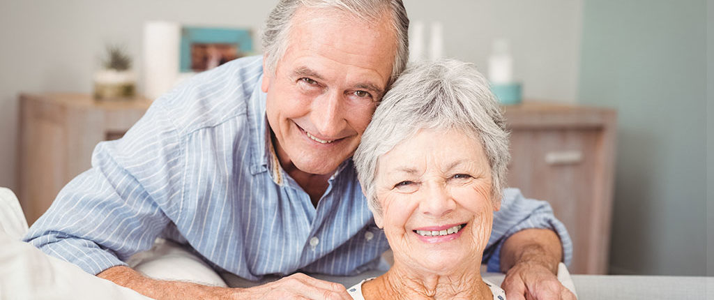 A man leans over a couch to pose with a woman who is seated, his hands are on her shoulders