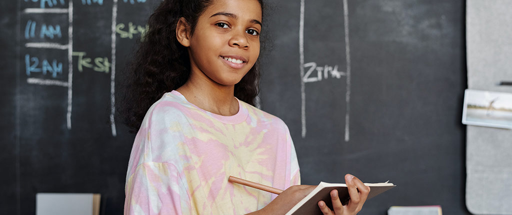 A young girl writes on a clipboard while standing in front of a blackboard