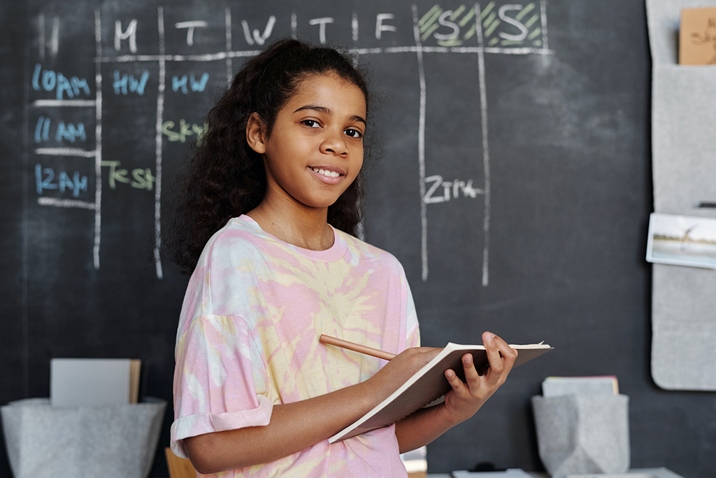 A young girl writes on a clipboard while standing in front of a blackboard