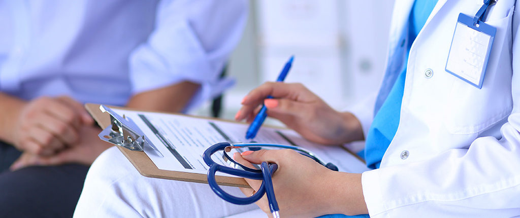 A woman holds a clipboard, stethoscope and pen. A man sits in the back