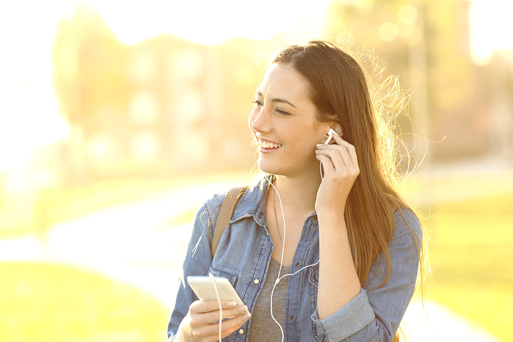 A smiling woman in a sunny park adjusts her left ear phone  