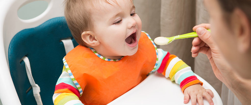 A baby in a white high chair, mouth open as an adult in the foreground feeds them from a green spoon