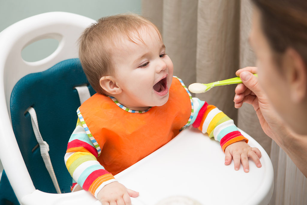 A baby in a white high chair, mouth open as an adult in the foreground feeds them from a green spoon