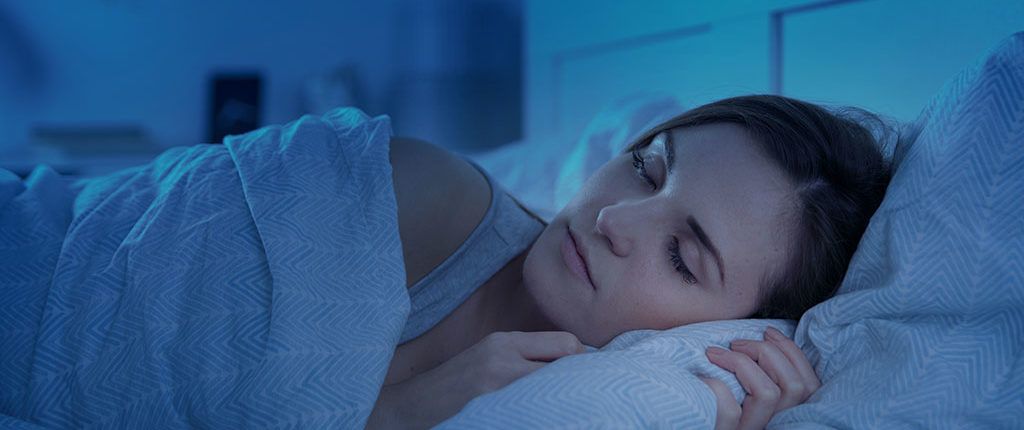 Light-skinned woman with dark hair sleeps peacefully under the covers in her bed