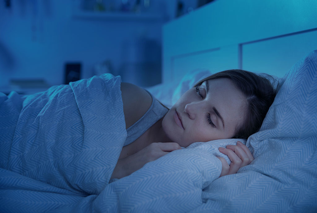 Light-skinned woman with dark hair sleeps peacefully under the covers in her bed