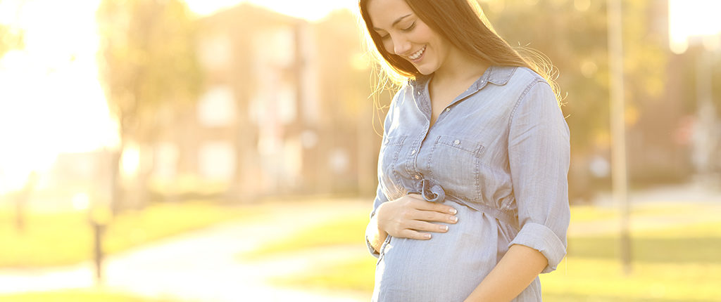 A pregnant woman in a blue shirt holds her belly while standing in a park
