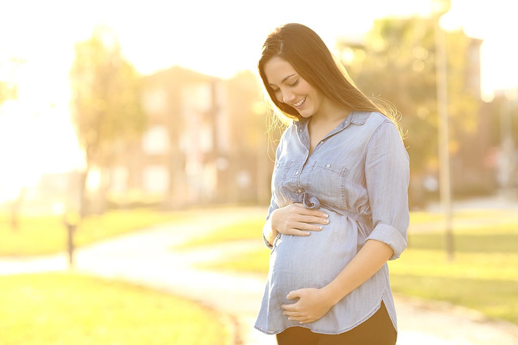 A pregnant woman in a blue shirt holds her belly while standing in a park