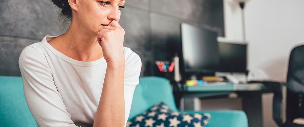 Light-skinned woman sits on a couch, fist propped against mouth as she looks to the side in worry