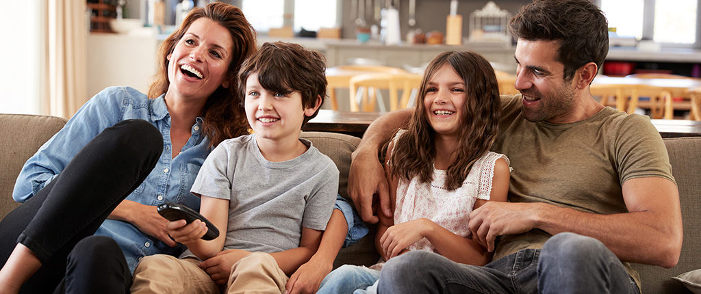 A family of four, a mother, son, daughter, and father, sits on a couch. The son holds a TV remote