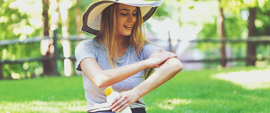 Pale-skinned woman in a sun hat and summer clothing sits on grass, rubbing sunscreen on her arm