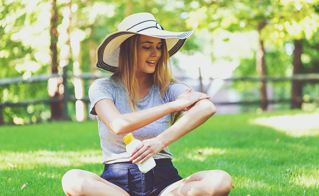 Pale-skinned woman in a sun hat and summer clothing sits on grass, rubbing sunscreen on her arm