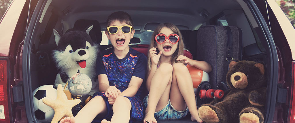 Two young children sit in the back of a liftgate trunk with toys and suitcases, yelling excitedly