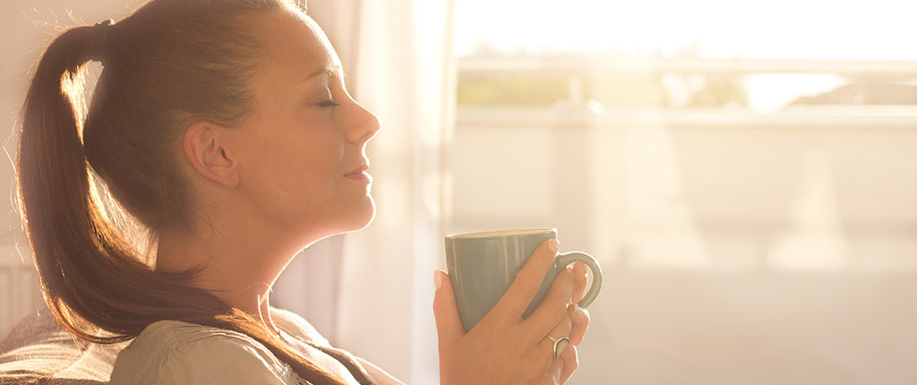 A woman with her eyes closed sits on a couch by a window holding a coffee cup