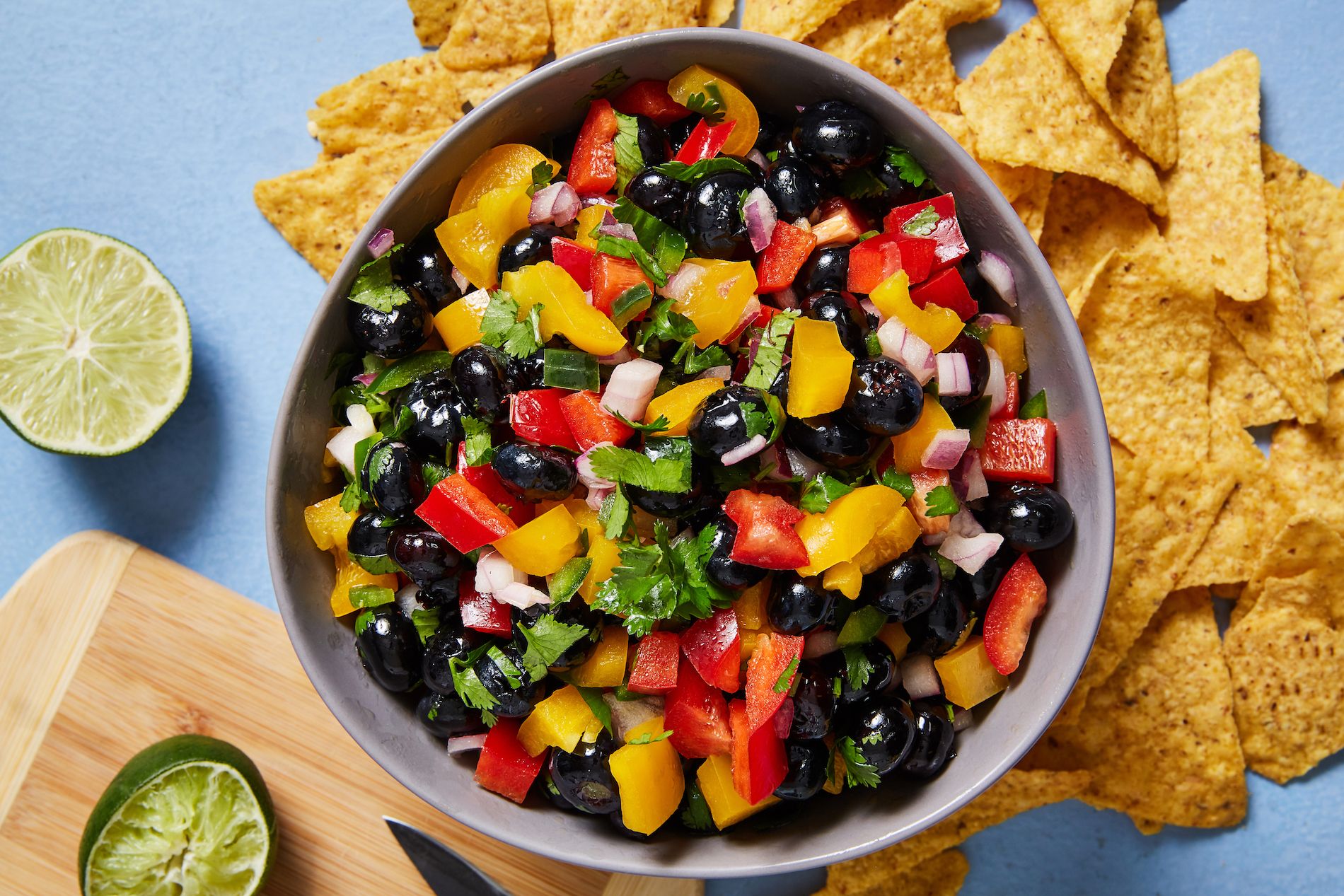 A bowl of salsa with beans, peppers, cilantro, and onions is surrounded by tortilla chips and limes