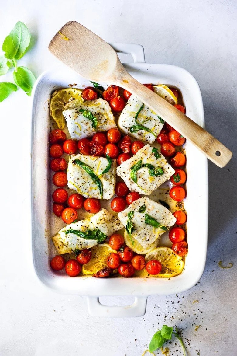 Top-down of a baked cod, cooked cherry tomatoes, and lemon slices in a white casserole dish