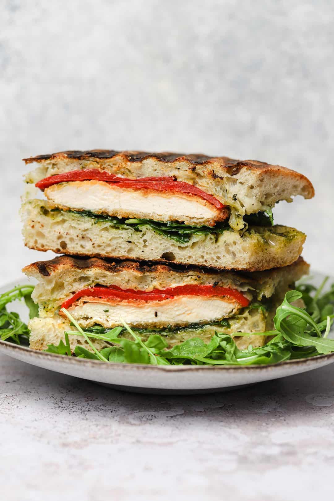 Close-up of two stacked halves of a chicken panini with tomato, greens, and pesto on a plate