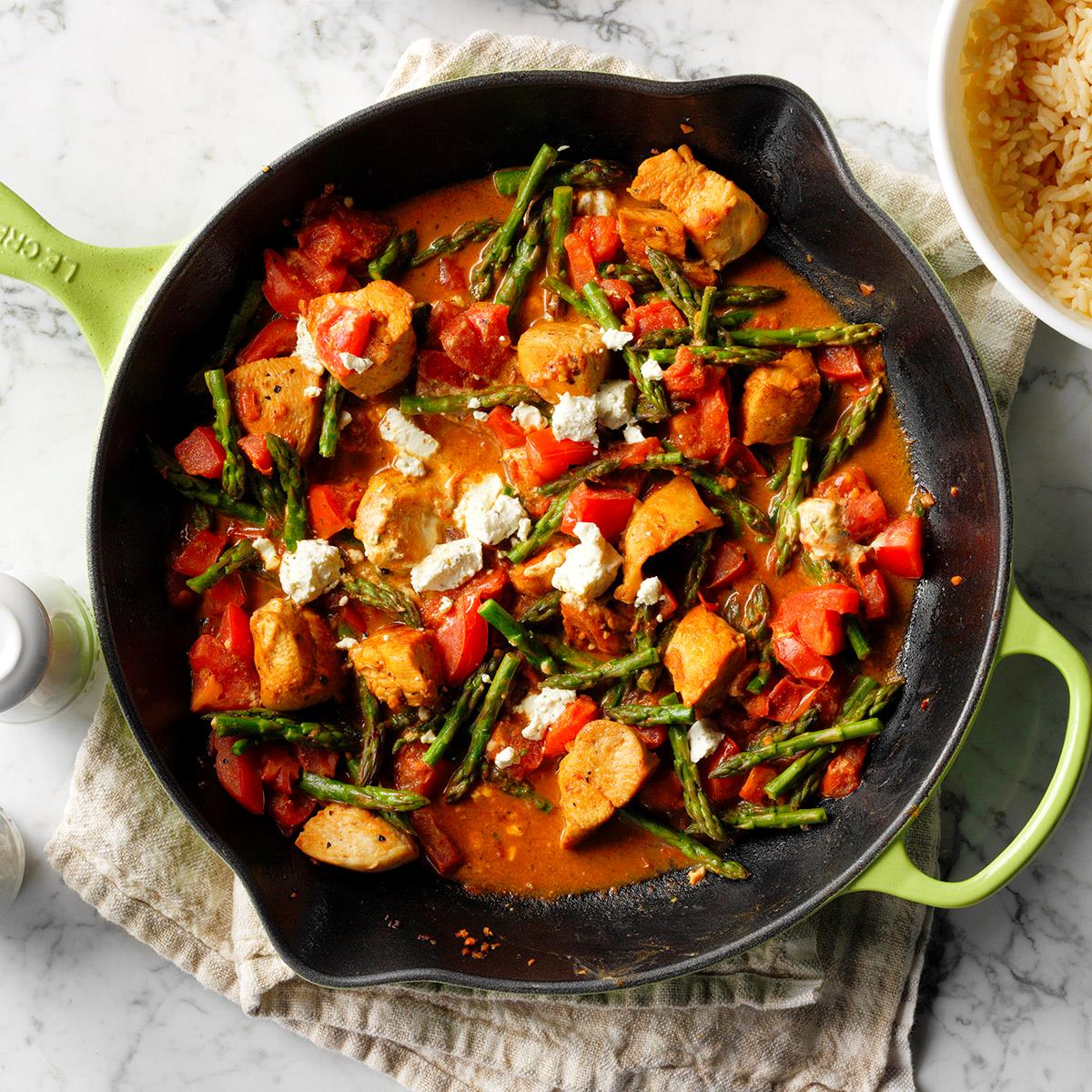 Top-down of a cast iron skillet with chicken, goat cheese, tomato, and asparagus in a red sauce