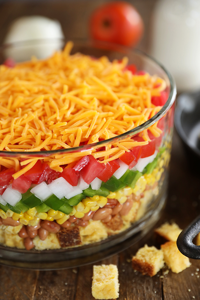 Glass bowl of cornbread salad, cornbread, beans, corn, peppers, onion, tomato, topped with cheese