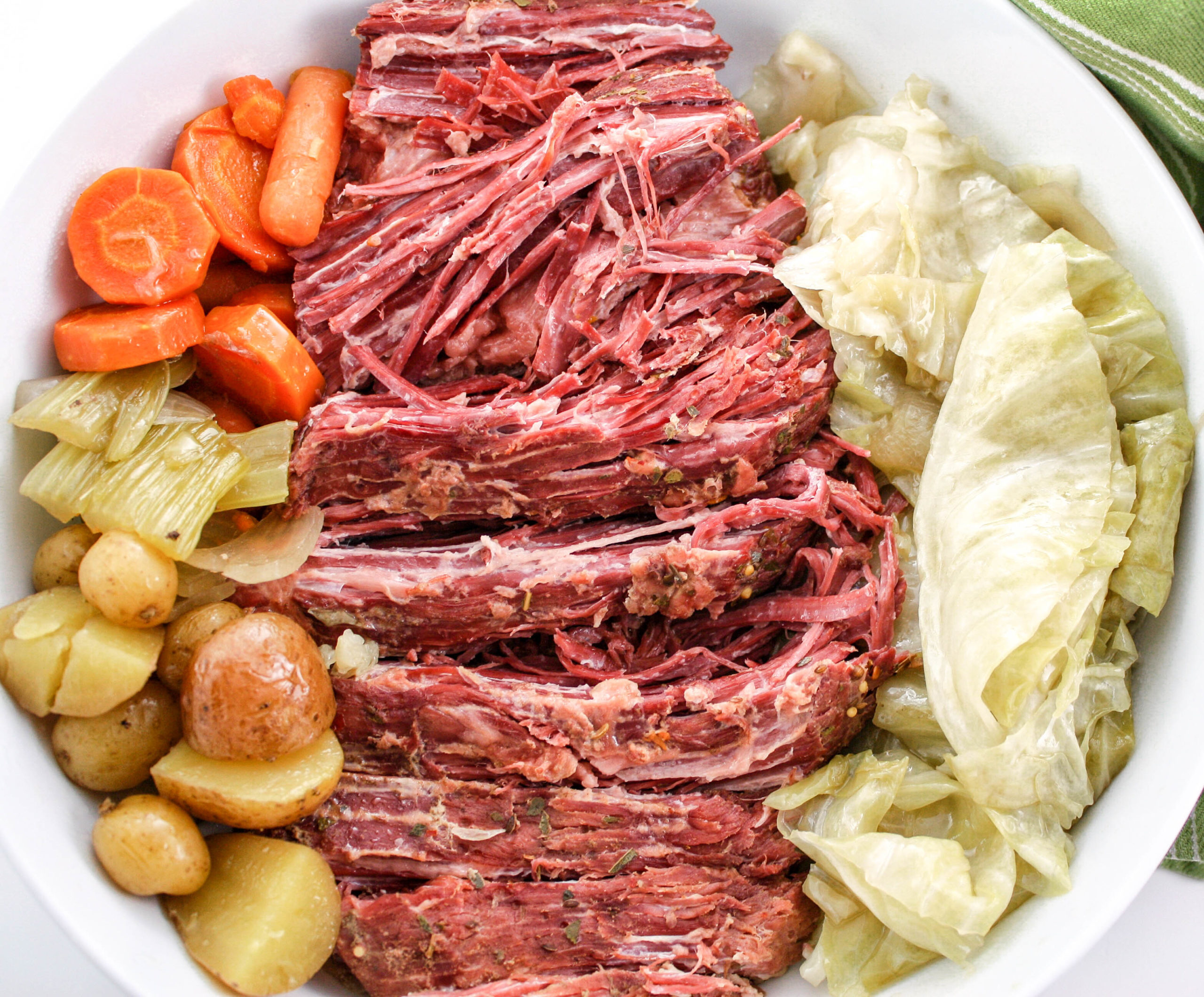 White bowl of corned beef surrounded by baby potatoes, cabbage, celery, and carrots