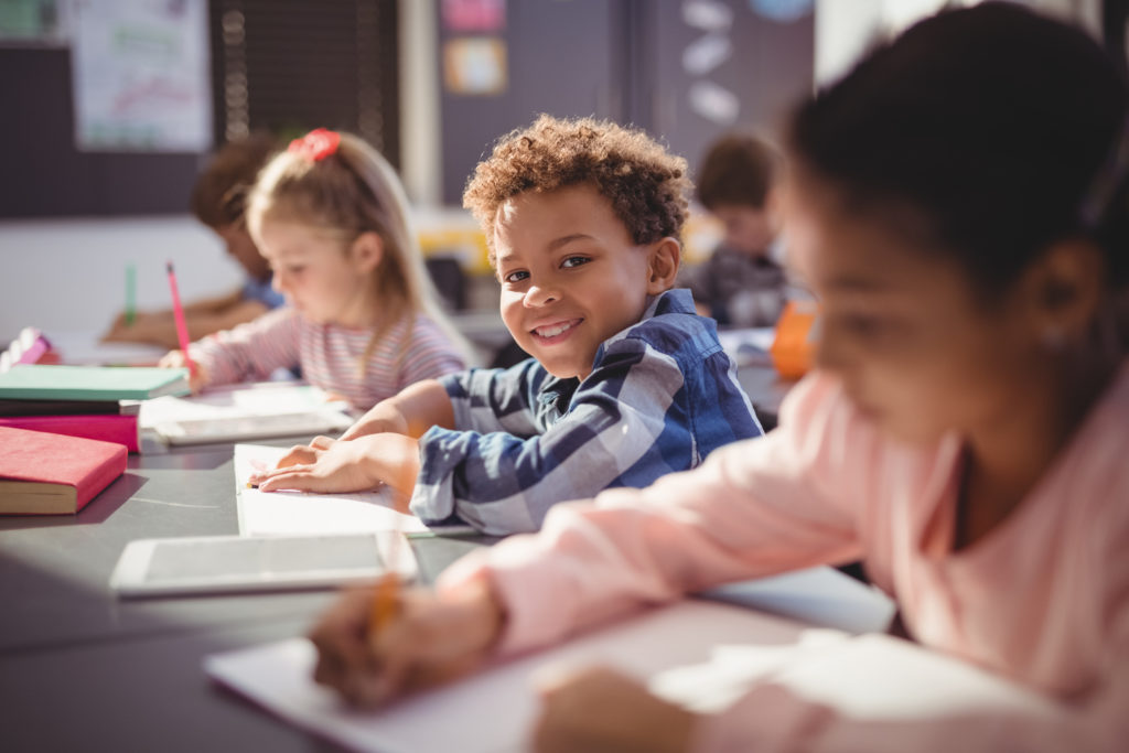 Child in class, smiling at camera as other students complete their workbooks. 