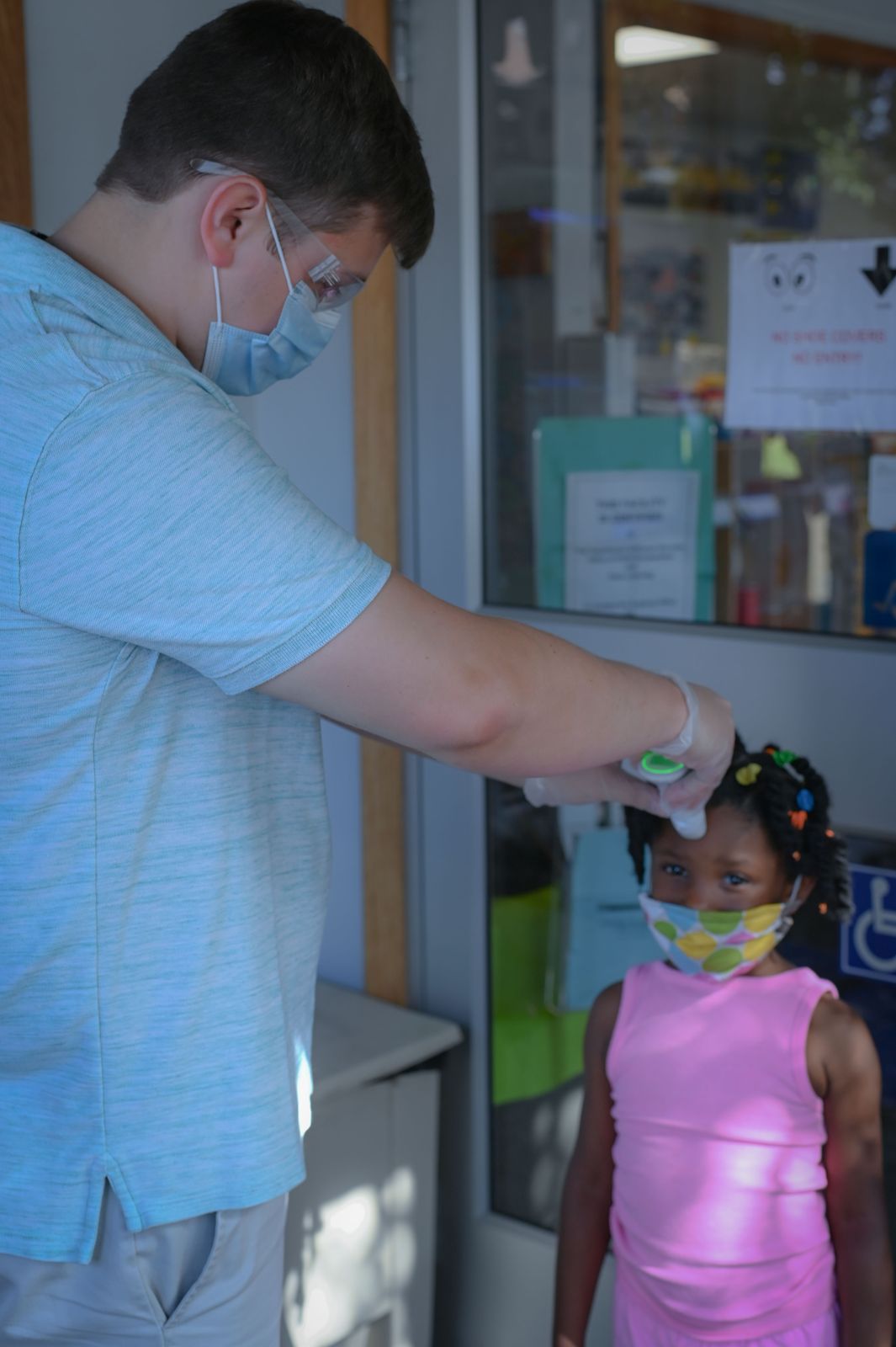 A man wearing a surgical mask uses a forehead thermometer on a young girl