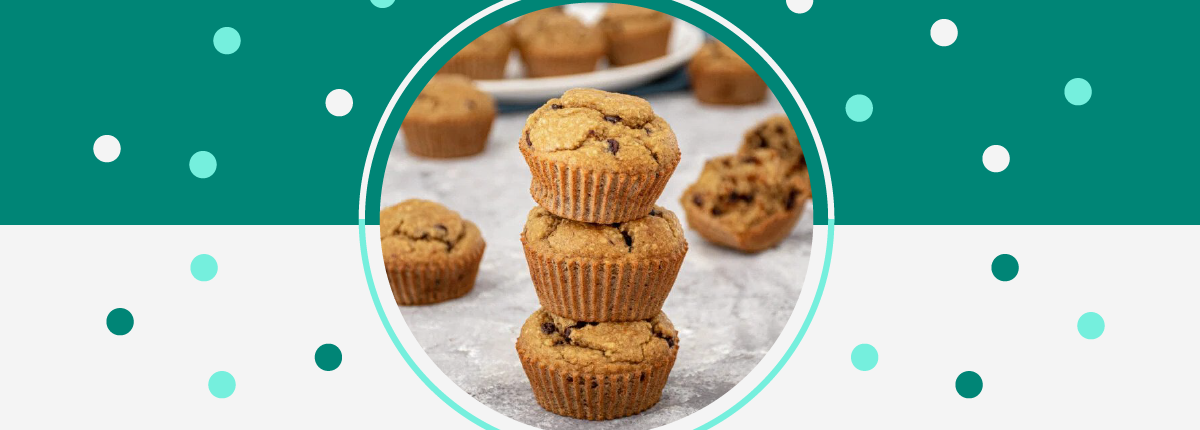 Three banana chocolate chip muffins sit stacked on a table with additional muffins in the background