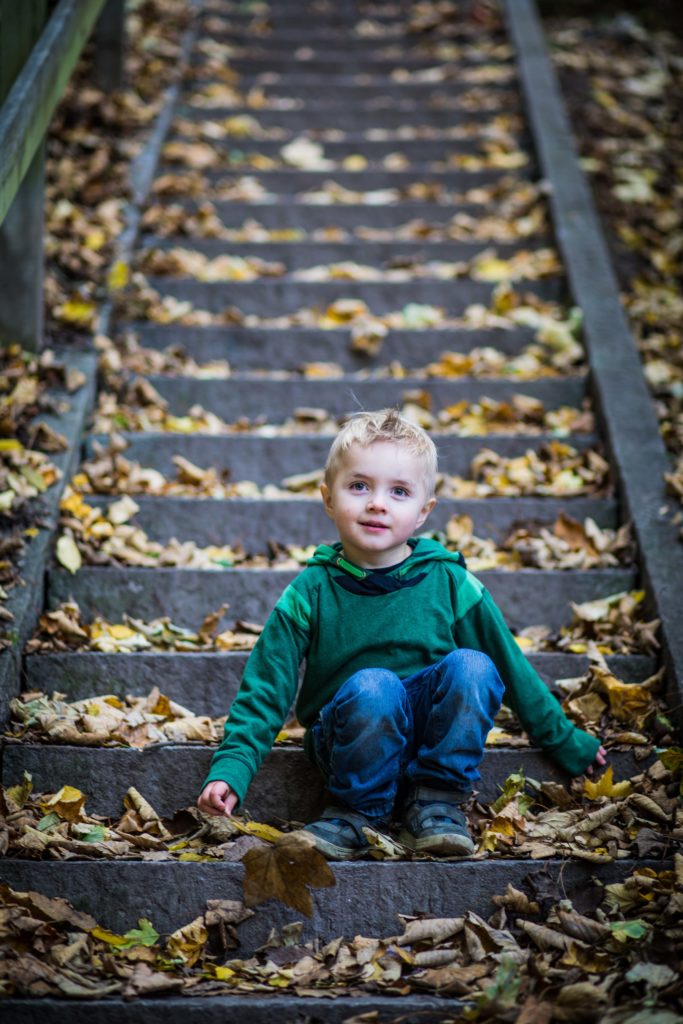 A young, blond child in fall clothing sits at the bottom of stone steps covered in brown leaves