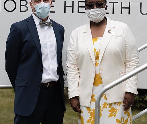 A man in a suit and surgical mask stands with a woman in a white dress, a blazer, and a cloth mask