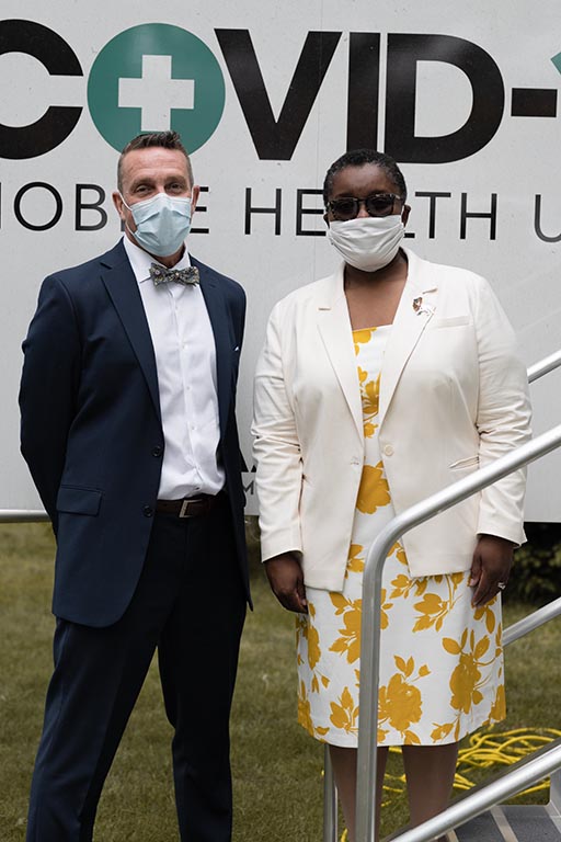 A man in a suit and surgical mask stands with a woman in a white dress, a blazer, and a cloth mask