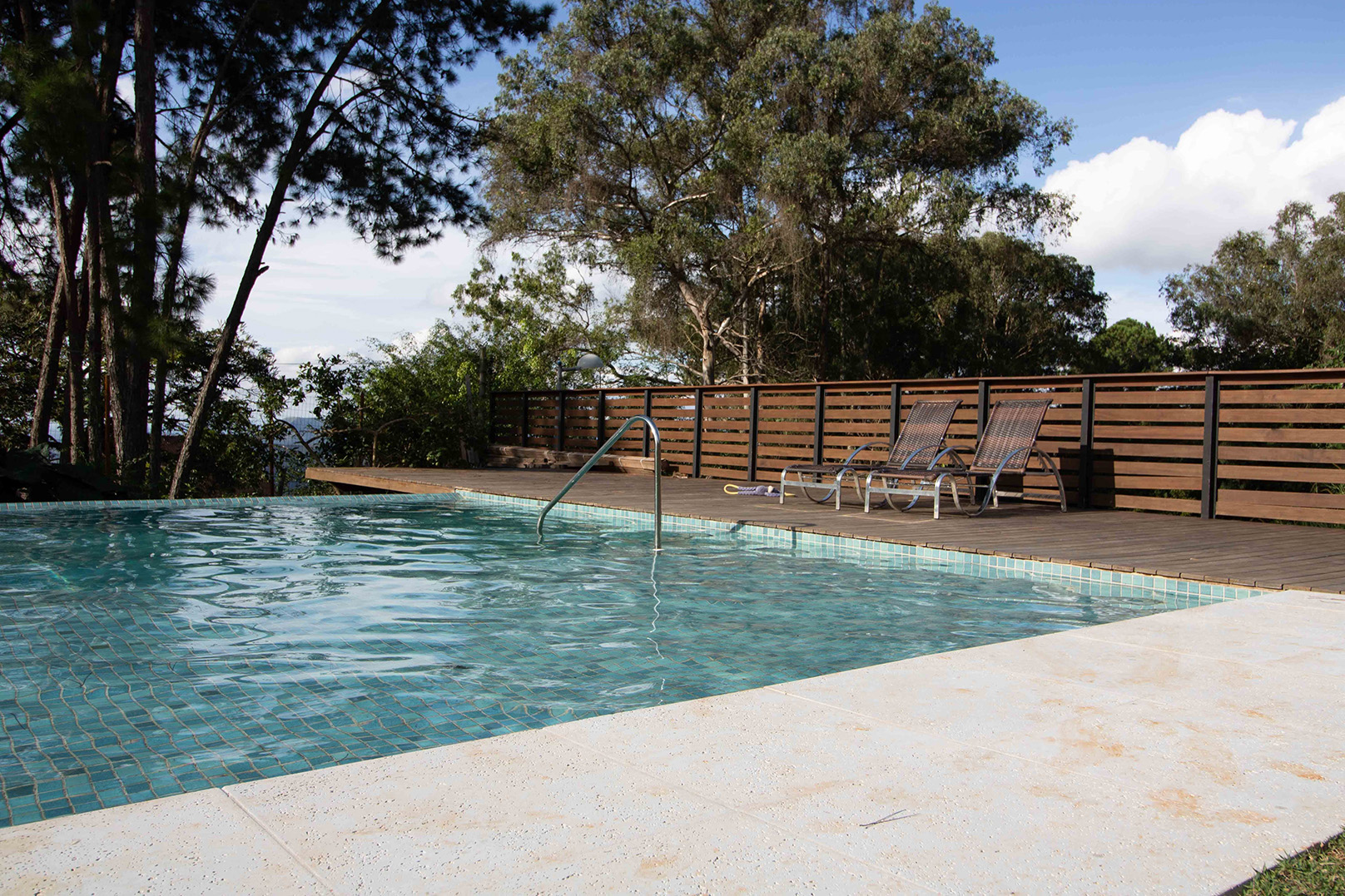 An at-home pool with a wooden deck and deck chairs to one side, trees and sky beyond it