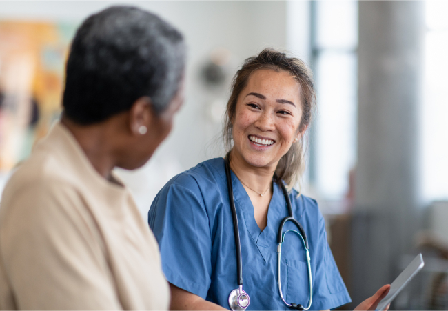 Angled view of a nurse in uniform holding a tablet and smiling at patient. 