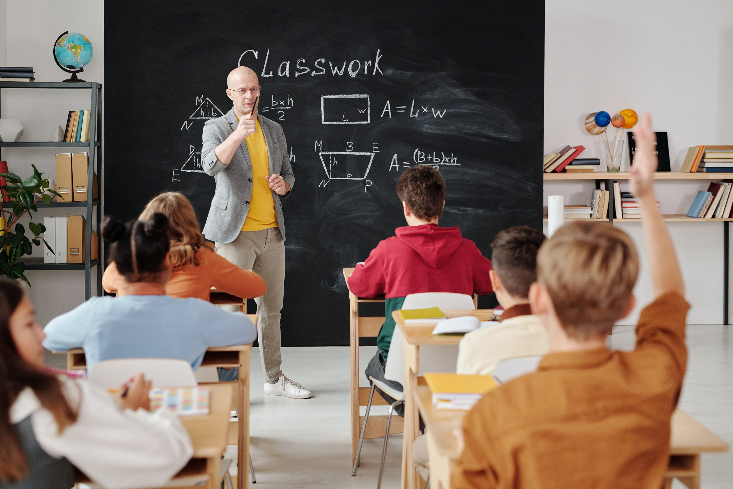 Teacher calls on a student in a classroom of adolescent children, a blackboard shows basic geometry