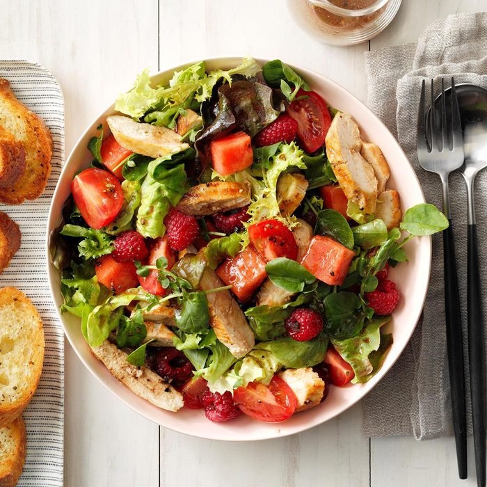Top-down of a salad with chicken, tomatoes, strawberries, and raspberries with bread and a drink