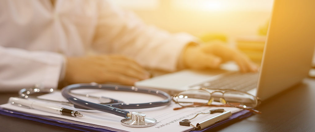 A clipboard with glasses, a stethoscope, and a pen. In the background is a laptop and a person