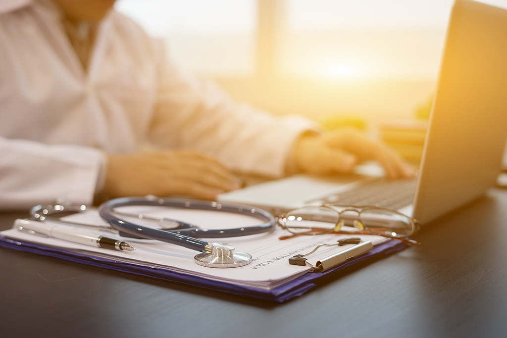 A clipboard with glasses, a stethoscope, and a pen. In the background is a laptop and a person